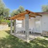 Bungalow bianco con tettoia in legno, veranda con tavolo e sedie. Circondato da erba e alberi.